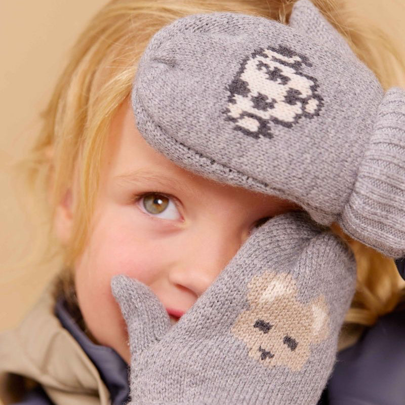 A close-up of a child wearing gray knit winter gloves. The glove has a beige bear face design, and the hat has a gray and white animal pattern, set against a warm beige background.