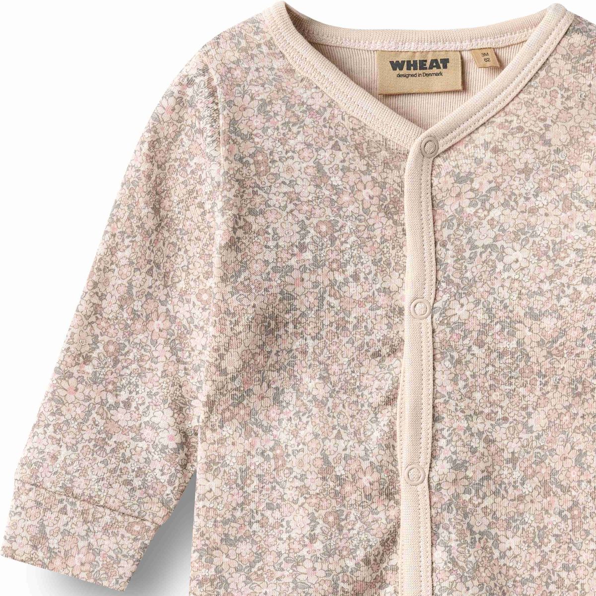 A close-up of a baby romper from the brand WHEAT, featuring a delicate pink and gray floral pattern on a light beige base, with a button-up front and ribbed trim.