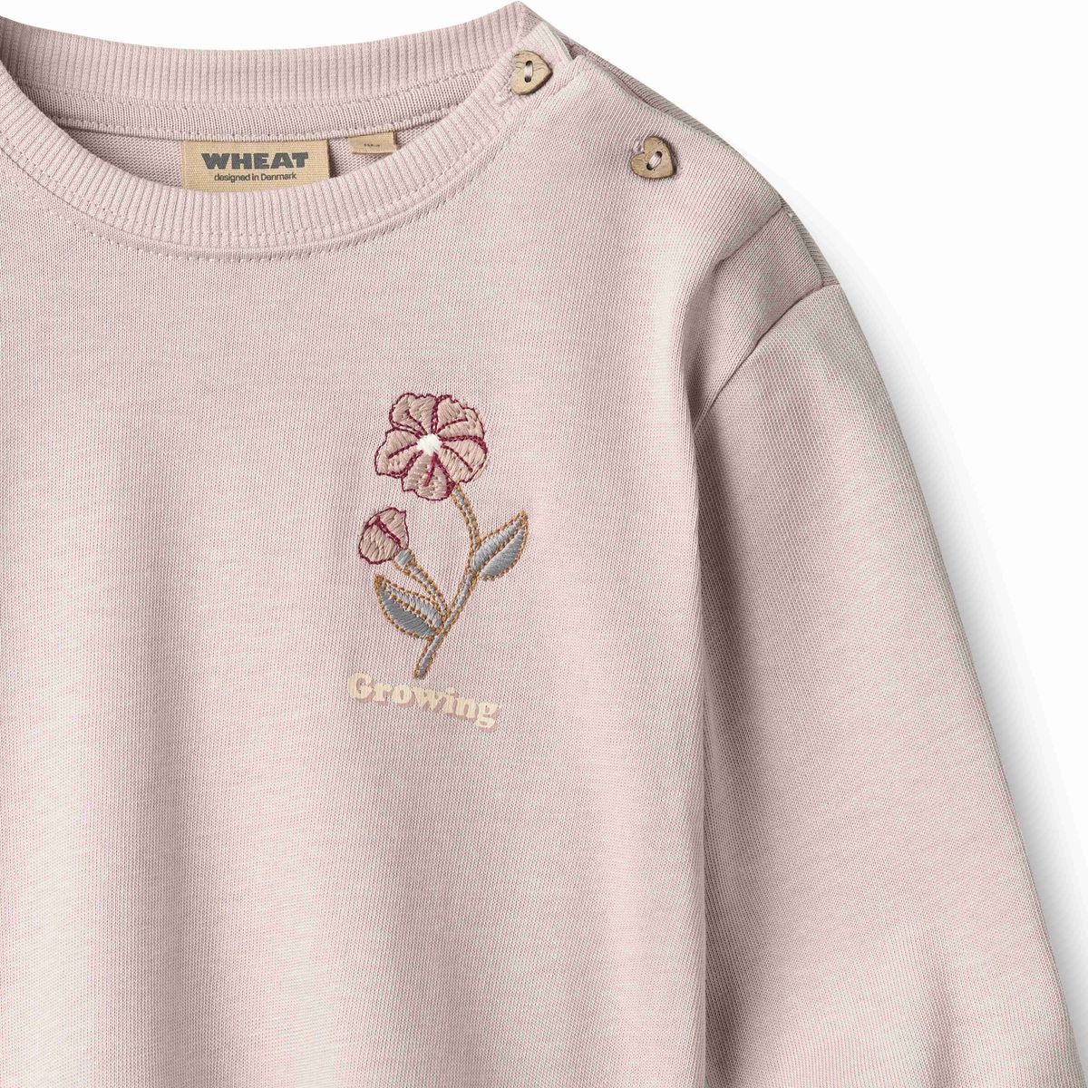 A close-up of a light pink crewneck sweatshirt with a floral embroidery on the chest and the word 'Growing' below it. The brand name 'WHEAT' is visible on the inner neck label.