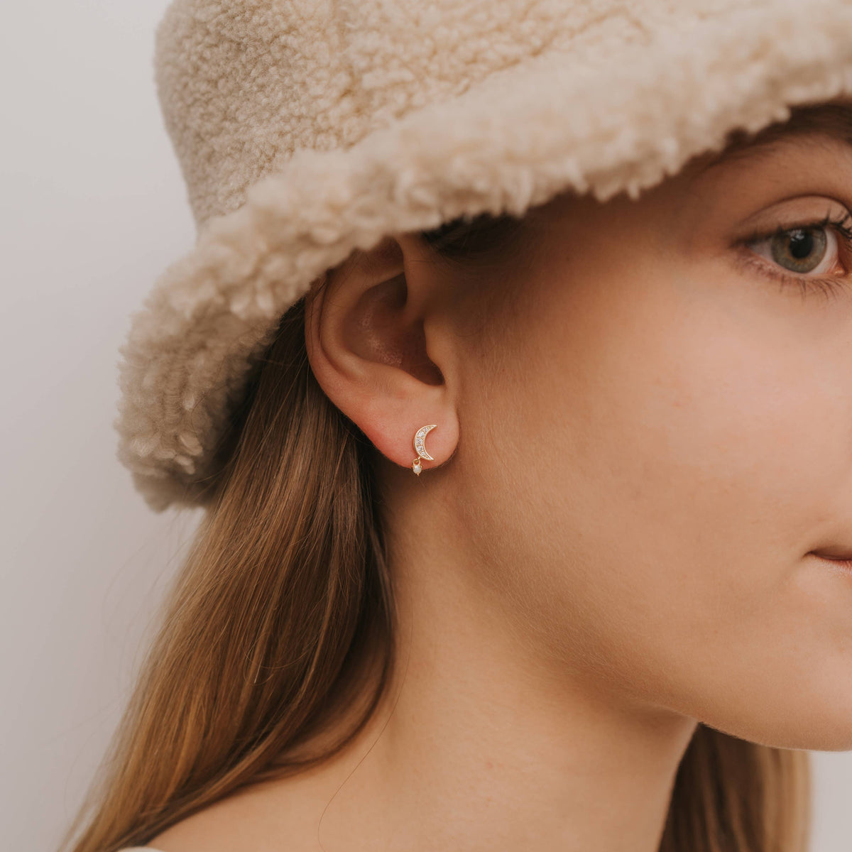 Close-up of a person wearing a beige hat and earring against a neutral background