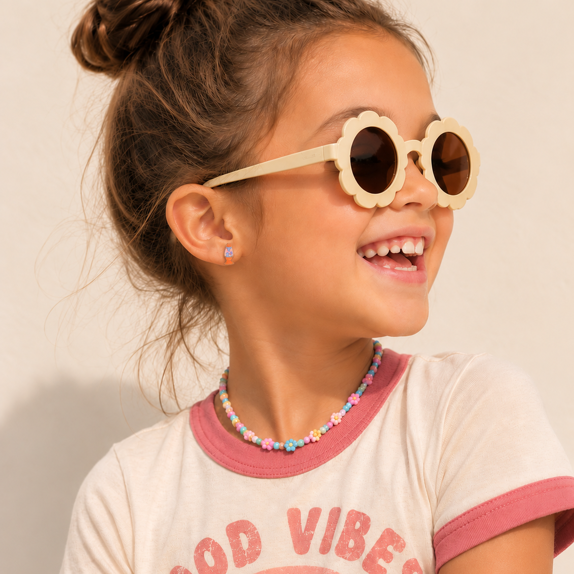 A young girl with her hair in a bun wearing scalloped round sunglasses with cream frames and dark lenses, a colorful beaded necklace, and a cream t-shirt with pink trim and a rainbow graphic that reads 'GOOD VIBES'.