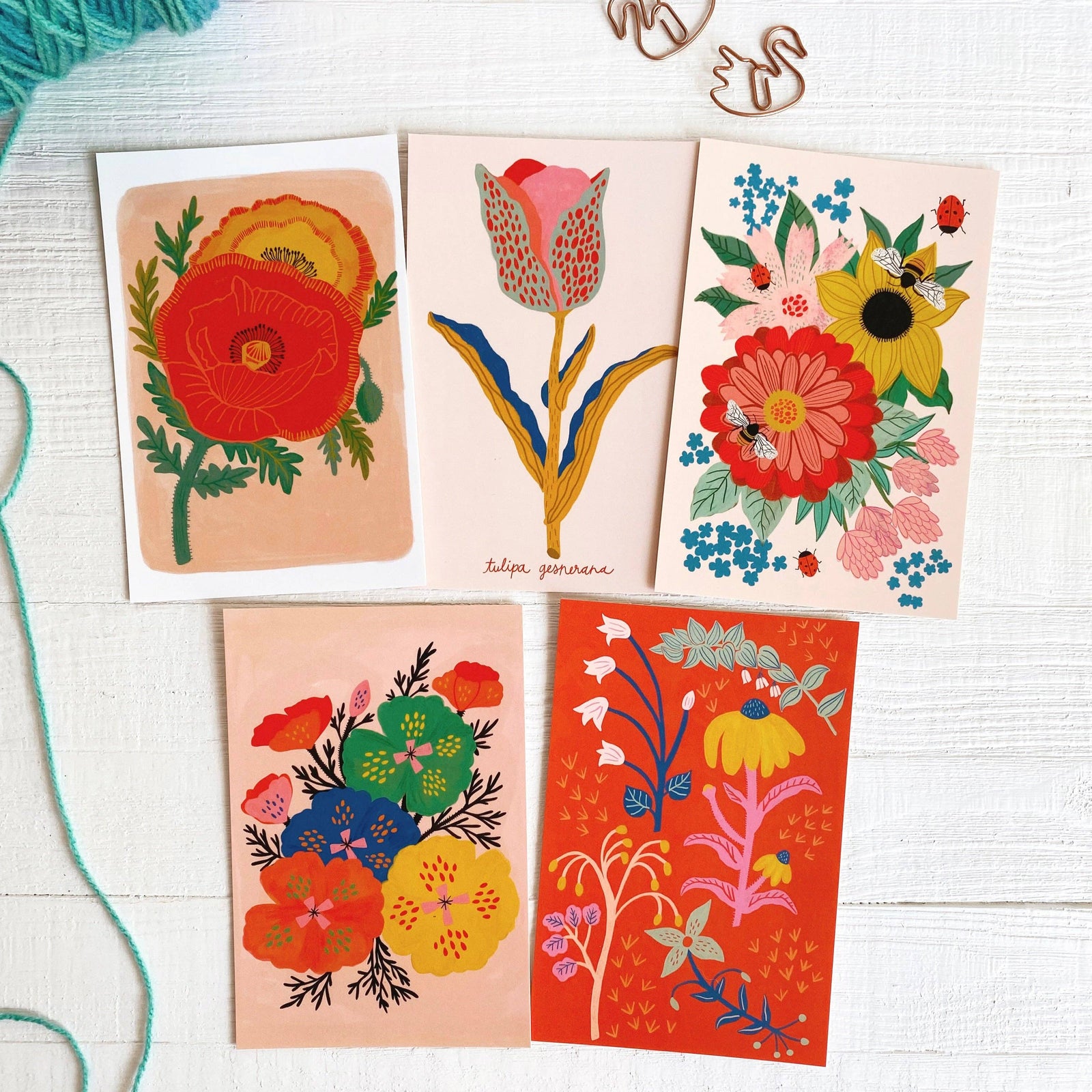 Five colorful floral greeting cards on a white surface with yarn and wooden swan decorations.