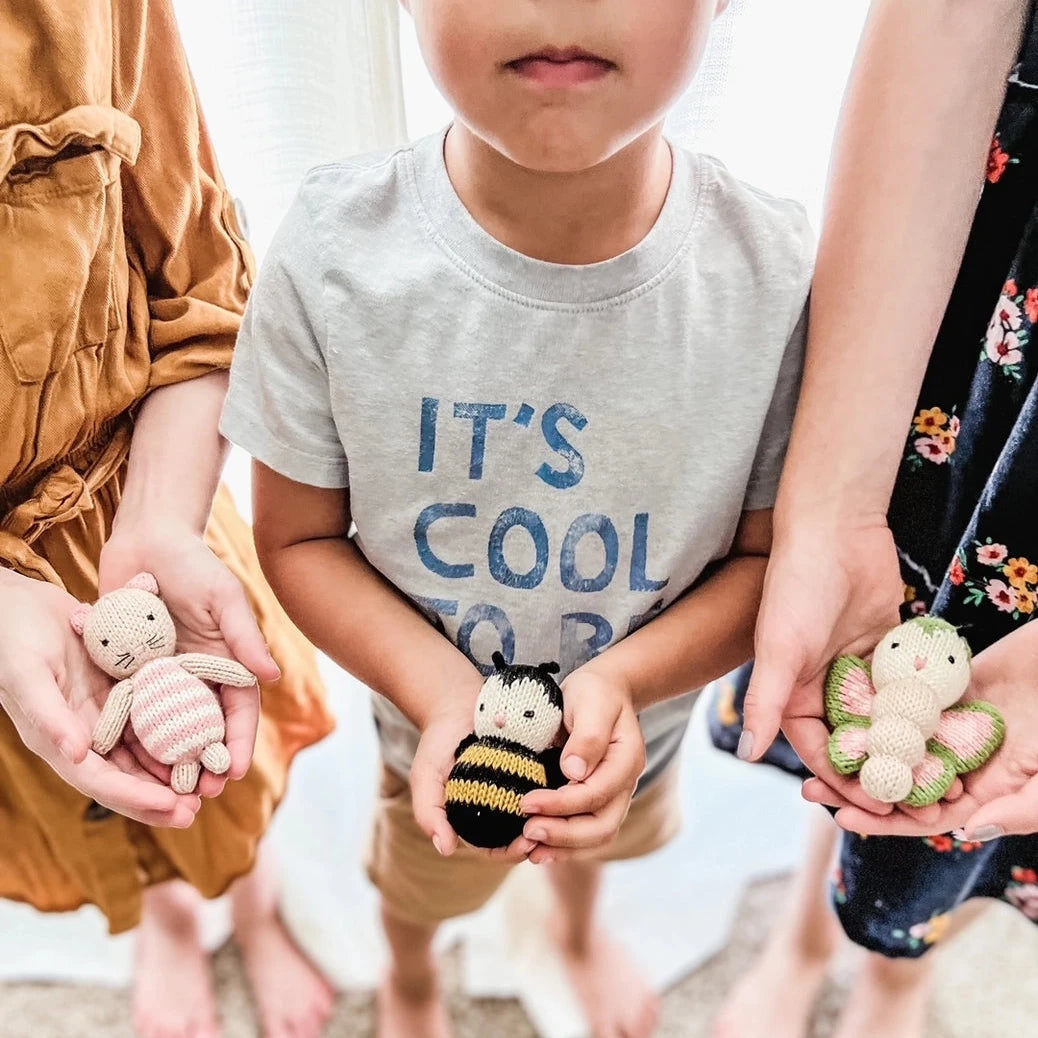 A child in a gray graphic t-shirt holding a small crocheted bumble toy, with other children holding small plush toys around them in a brightly lit indoor setting.