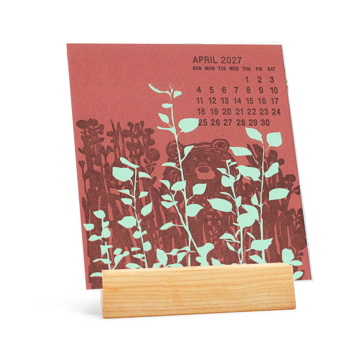 A vertical desk calendar page in terracotta red with a dark brown floral and plant illustration, plus a small April 2027 calendar grid at the top, displayed on a light natural wood stand against a white background.