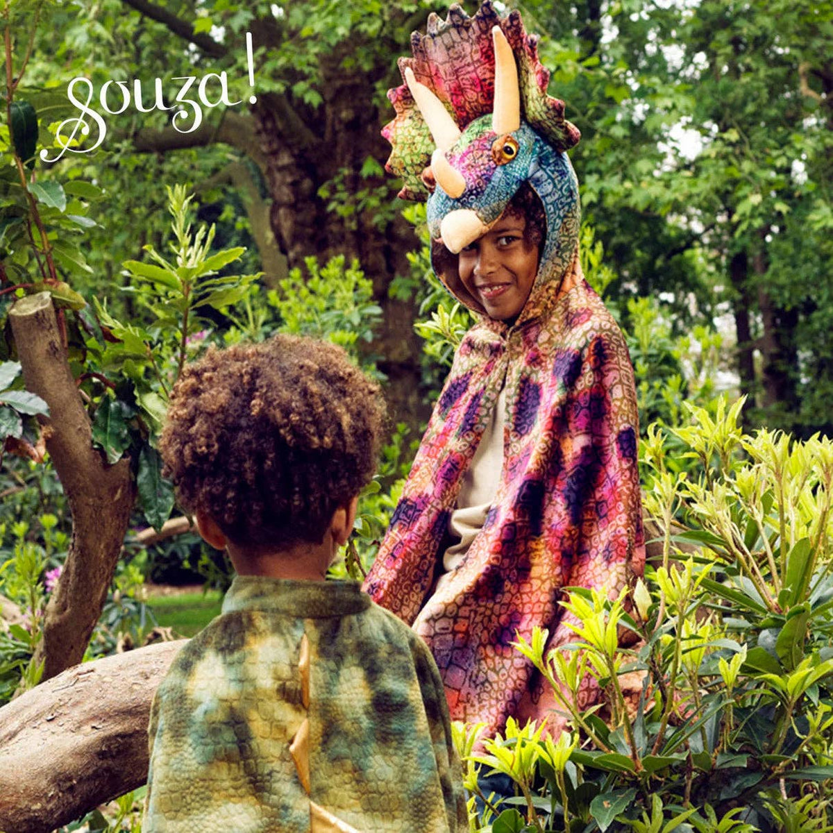 Two children in a forest setting, one wearing a dinosaur costume with a colorful cape.