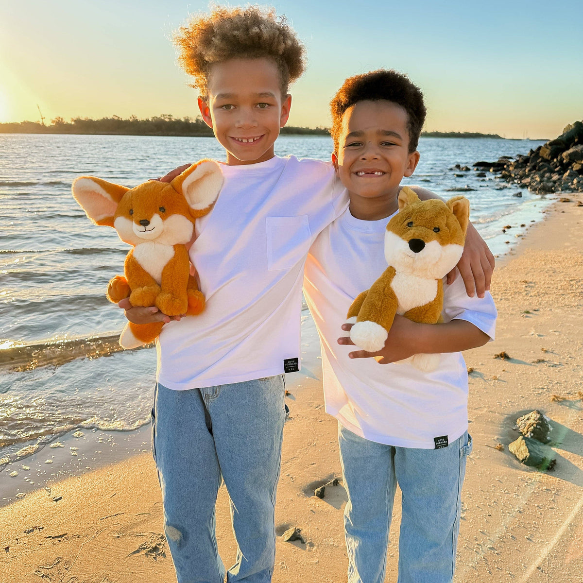 Two children holding stuffed animals on a beach at sunset