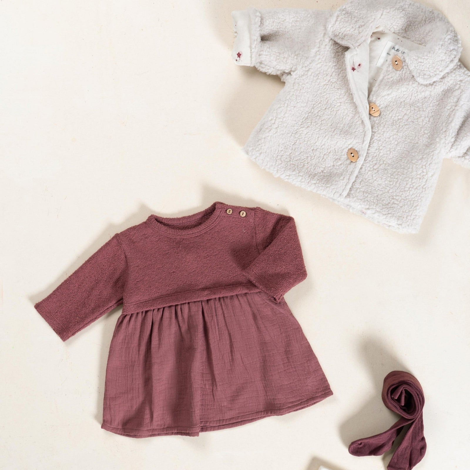 Children's clothing including a maroon dress, gray jacket, and purple tights on a light beige background.