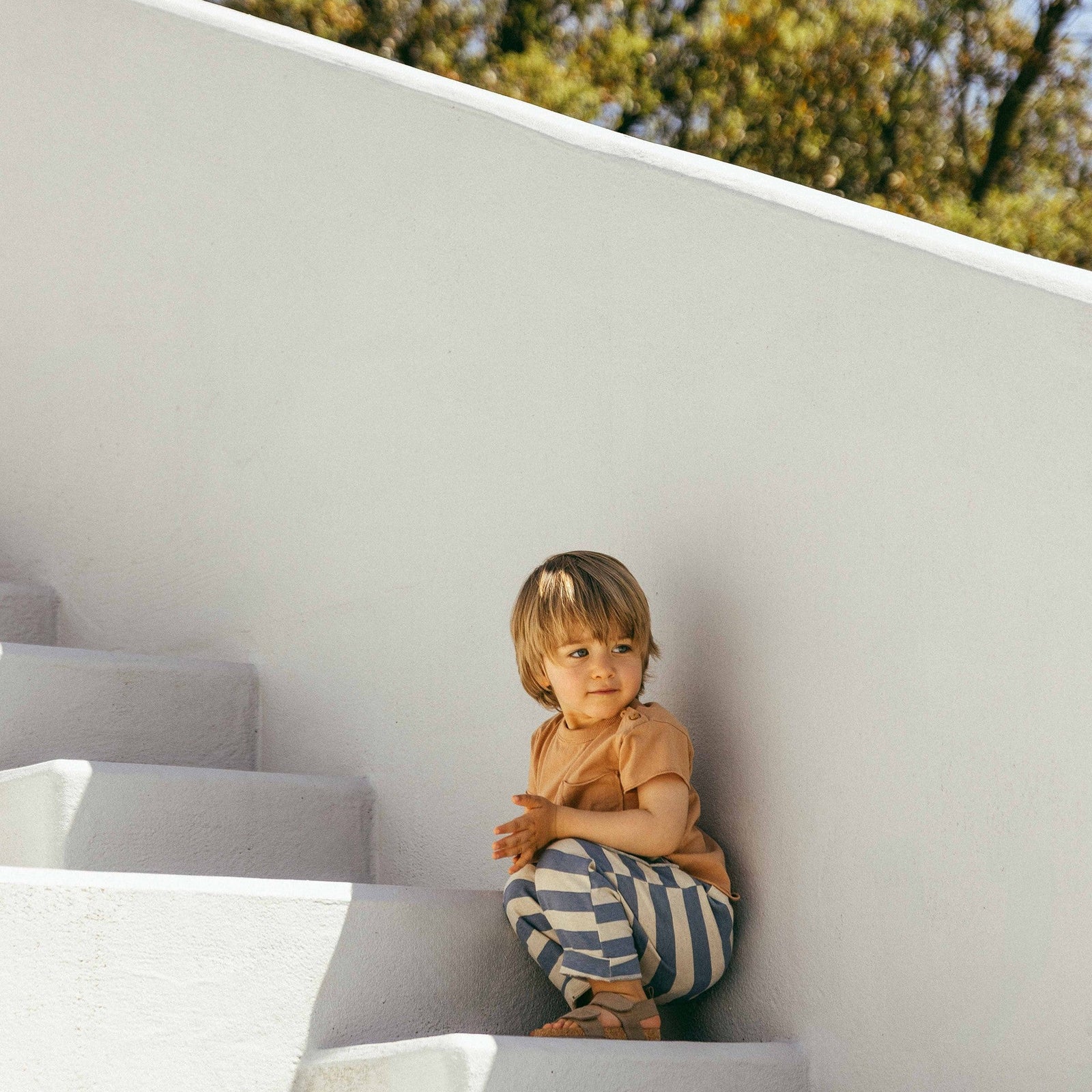 Child holding a red toy in a grassy area with a white wall and trees in the background