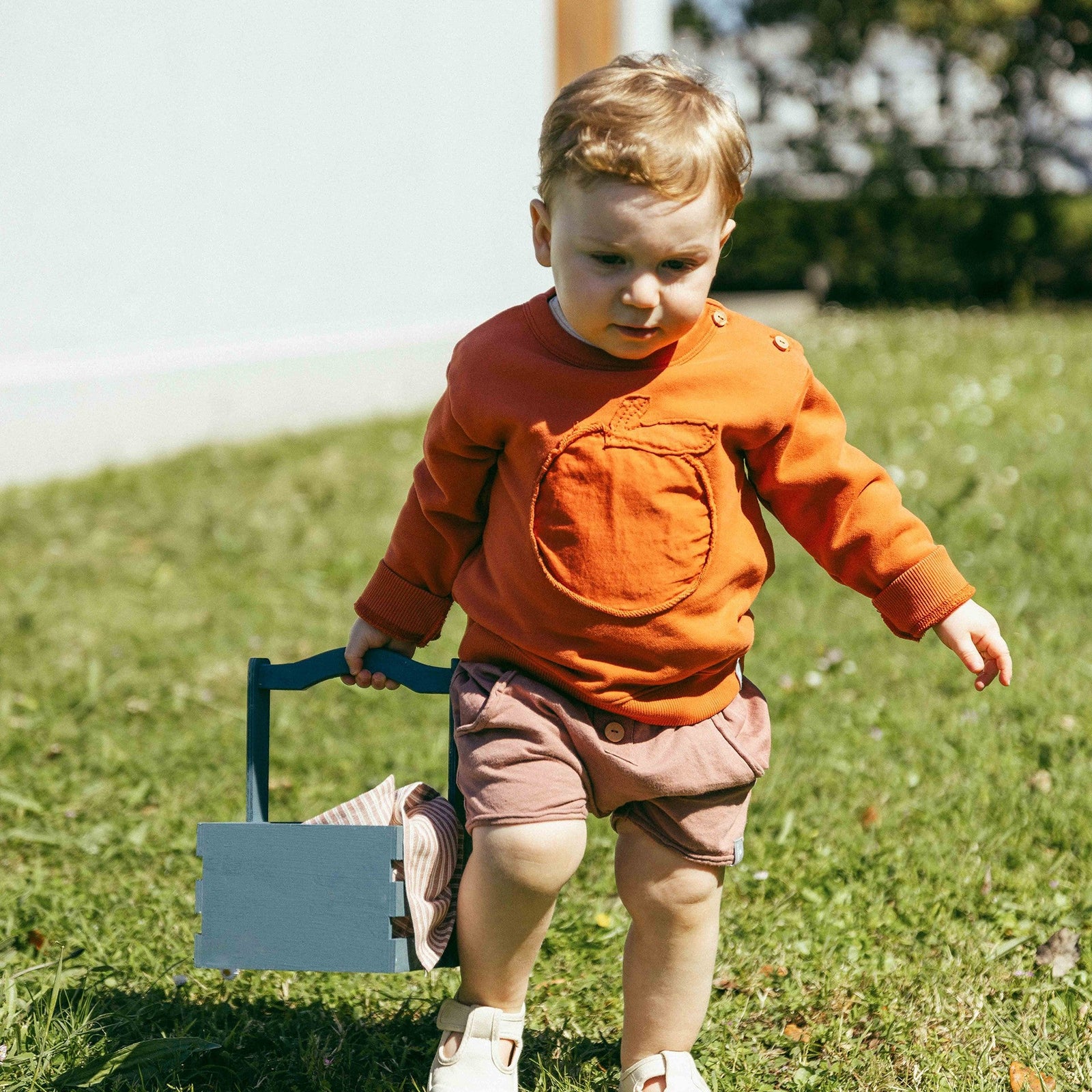 Child in an orange shirt and brown shorts walking outdoors with a toy shovel.