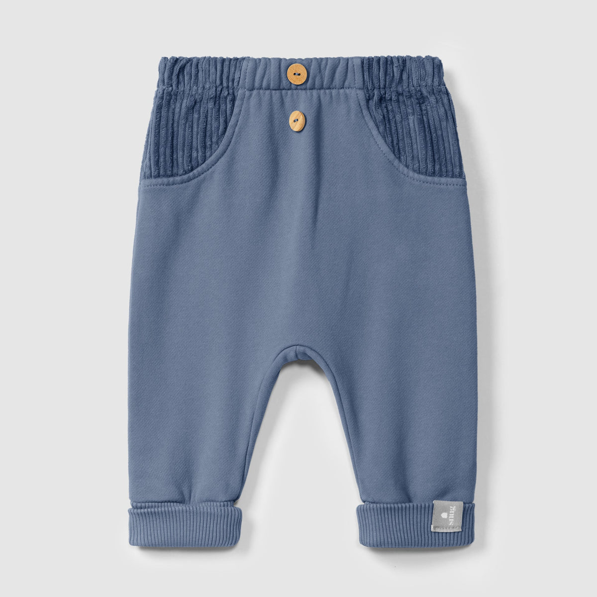 A pair of baby pants in dusty blue, featuring a textured elastic waistband with two wooden buttons, side pockets, and ribbed ankle cuffs, shown against a light gray background.