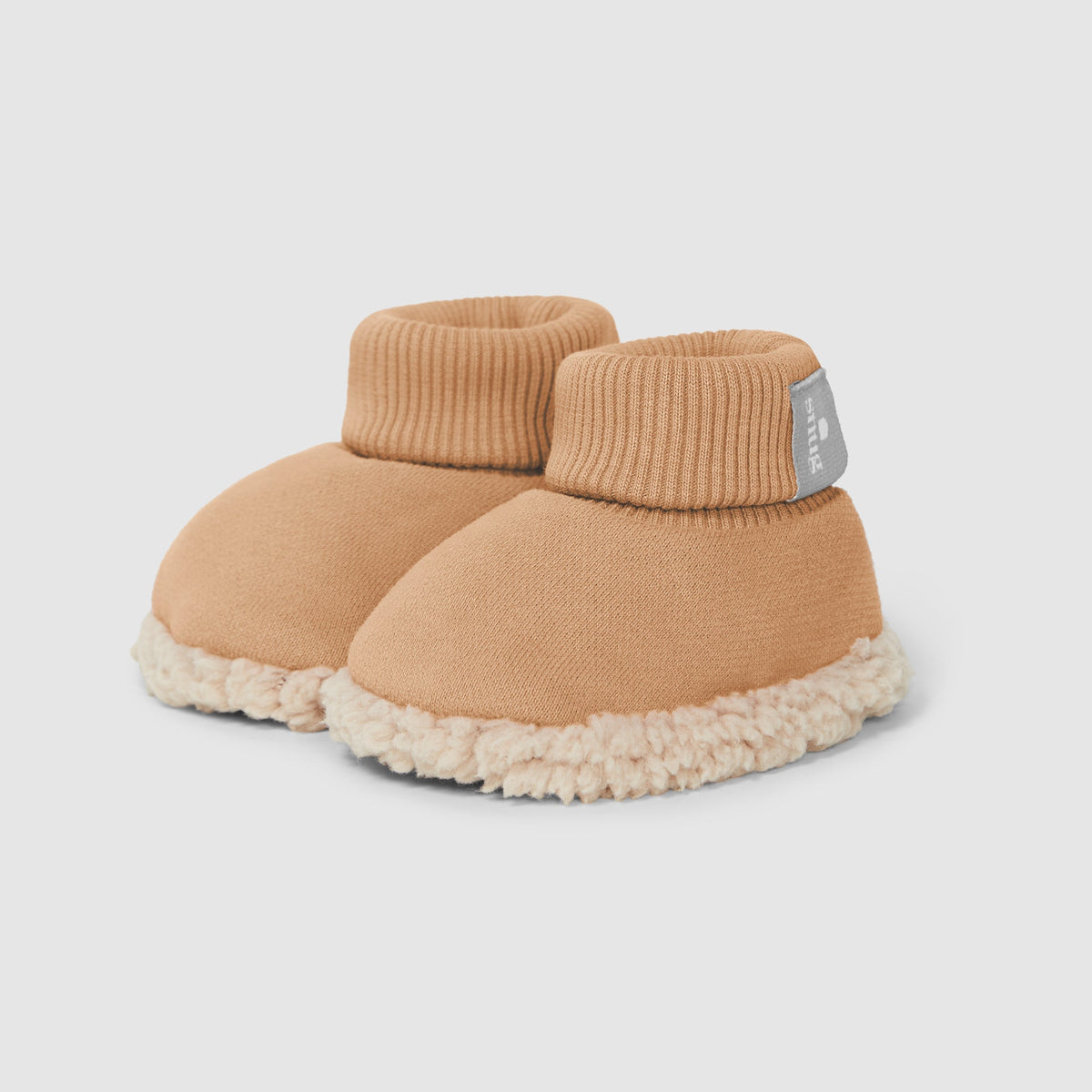 A pair of baby knit slipper boots in a warm camel tan color, featuring a soft cream sherpa fleece trim along the bottom edge and a small gray fabric brand tag on the side.