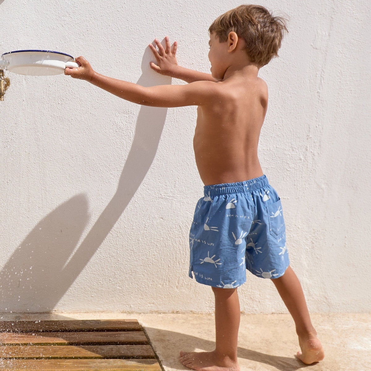 Child wearing blue swim shorts with a pattern, standing by a wall.