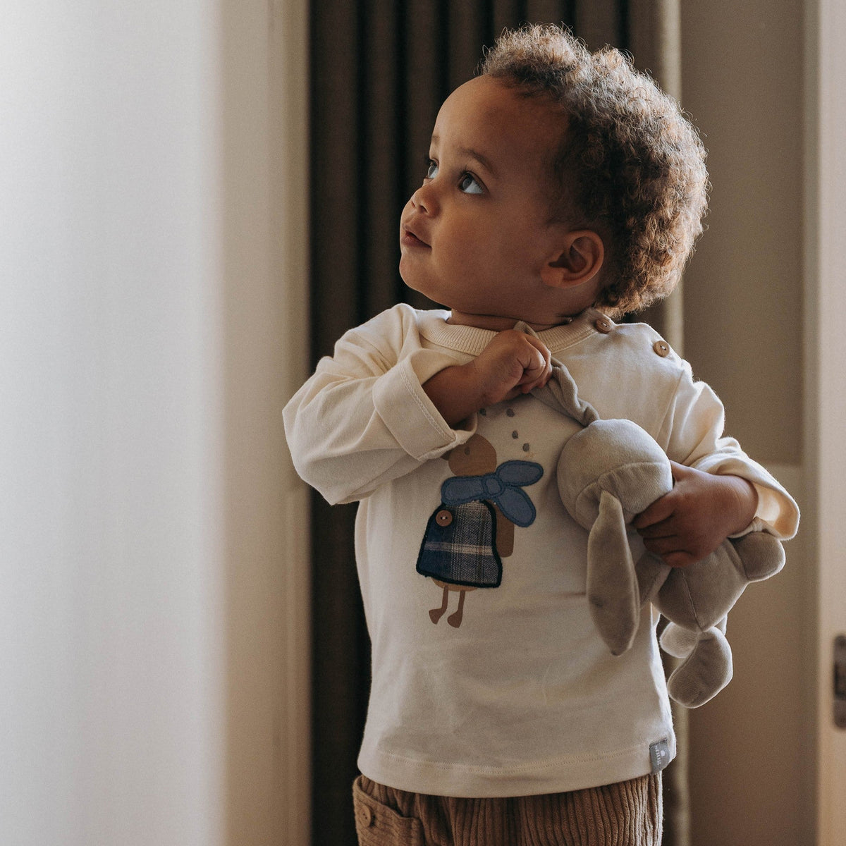 A toddler standing indoors near a window, wearing a cream long-sleeve shirt with a blue duck graphic and brown corduroy pants, while holding a gray plush toy.