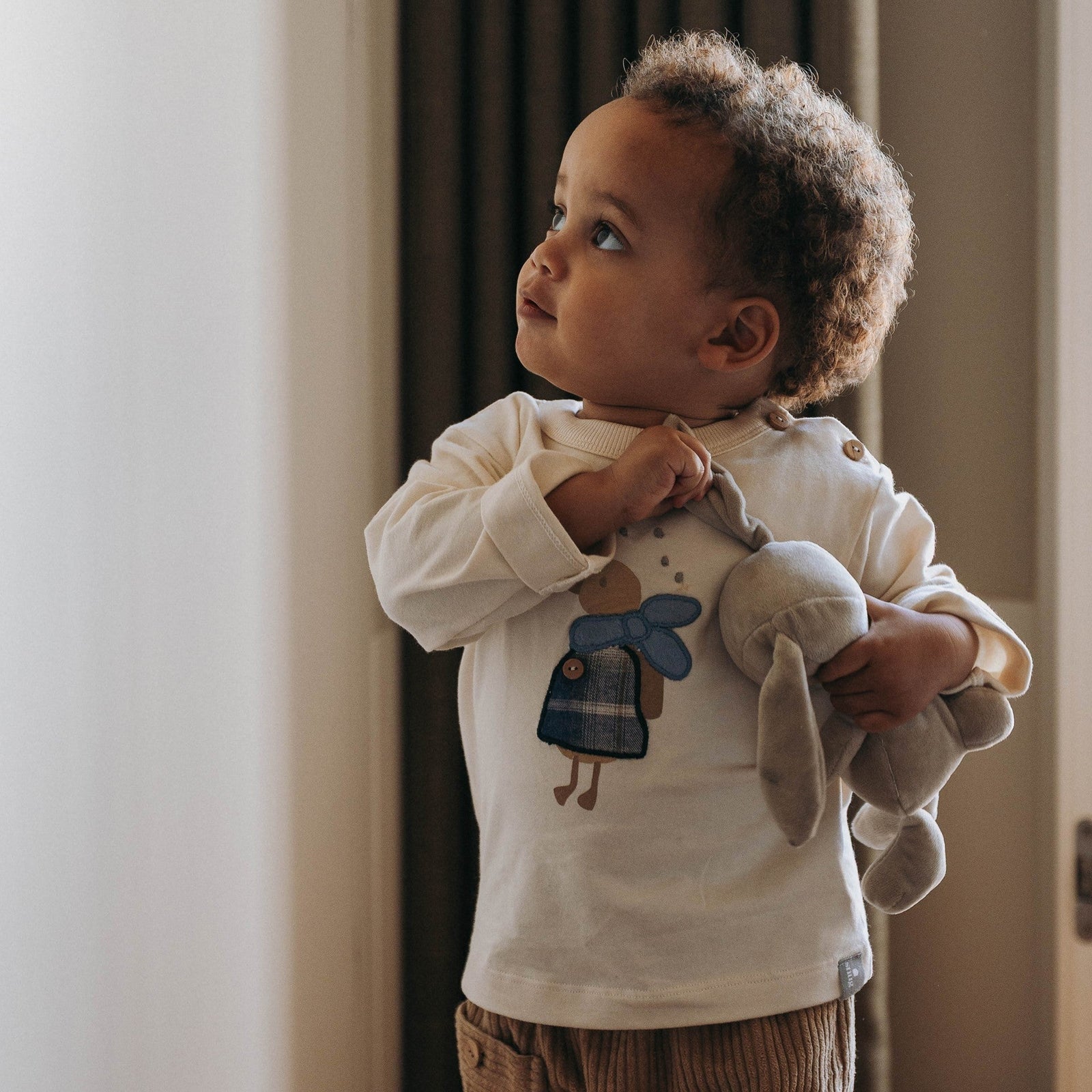 A toddler standing indoors near a window, wearing a cream long-sleeve shirt with a blue duck graphic and brown corduroy pants, while holding a gray plush toy.
