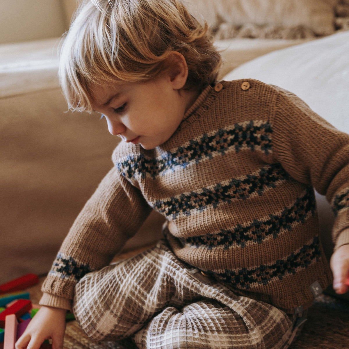 A young child sitting on a carpeted floor playing with colorful wooden blocks, wearing a brown knit sweater with dark patterned horizontal stripes and matching brown plaid pants.
