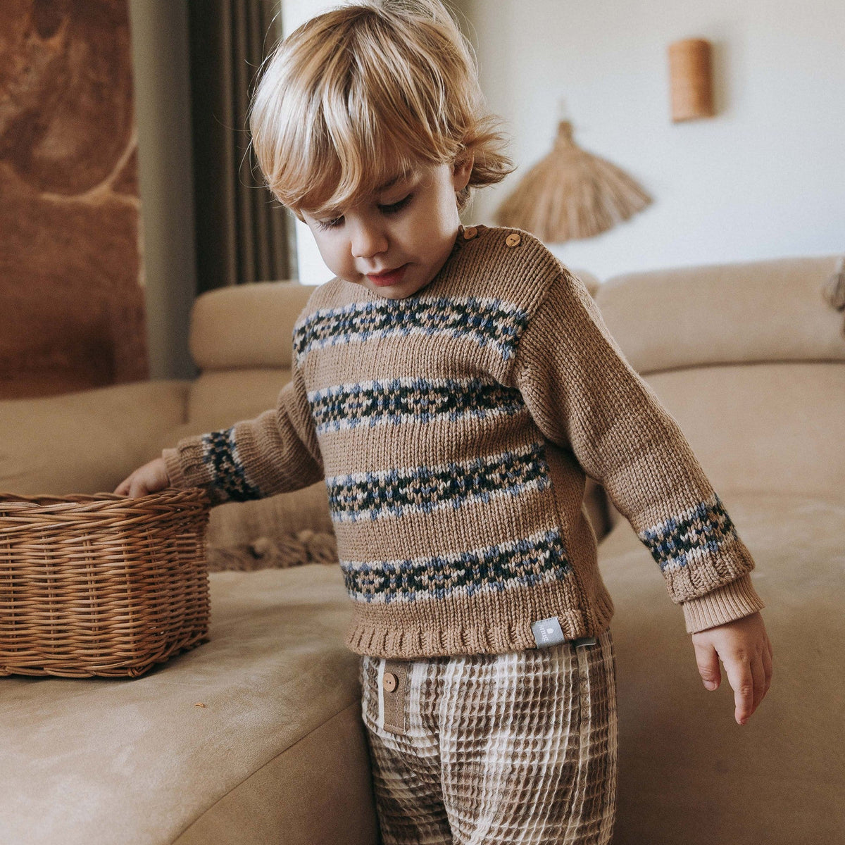 A young child wearing a beige knit sweater with dark blue and white geometric patterns and matching plaid pants, standing in a cozy living room next to a wicker basket.