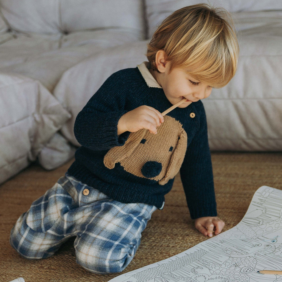 A young child with blonde hair kneels on a woven rug in front of a tufted sofa, wearing a navy knit sweater with a brown dog face patch and blue plaid pants, while coloring in a book.