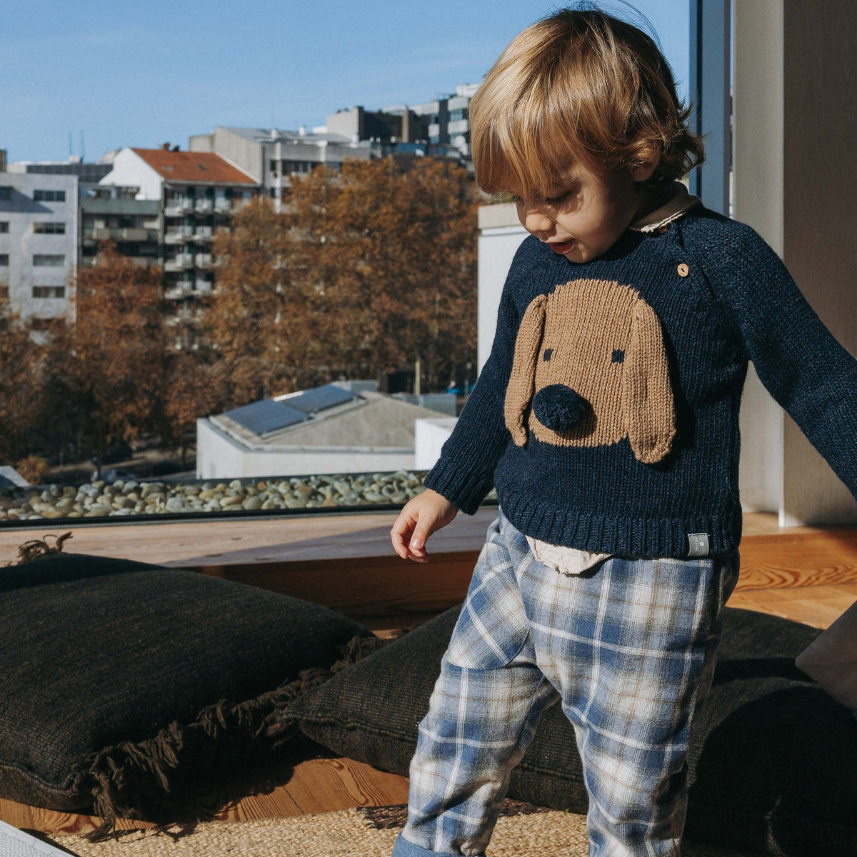A toddler stands indoors near a large window wearing a navy knit sweater with a tan dog face patch on the front, blue-and-white plaid pants, and dark socks. The room has a woven rug, dark cushions, and an outdoor city view with trees and buildings in the background.