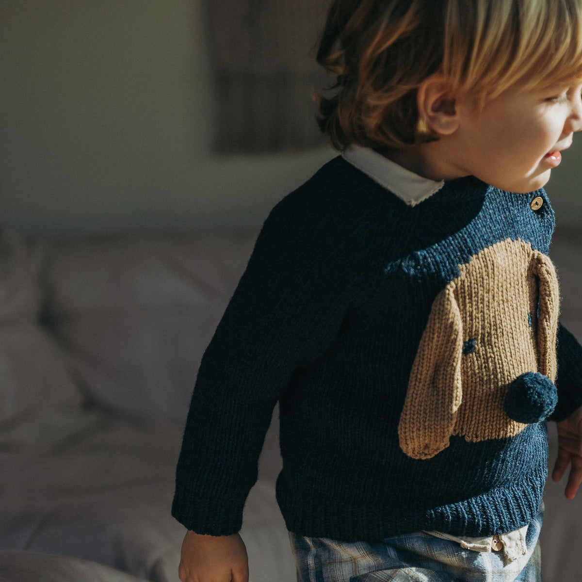 A toddler wearing a navy blue knit sweater with a large tan dog face applique on the front, paired with blue plaid pants, standing in a softly lit indoor room.