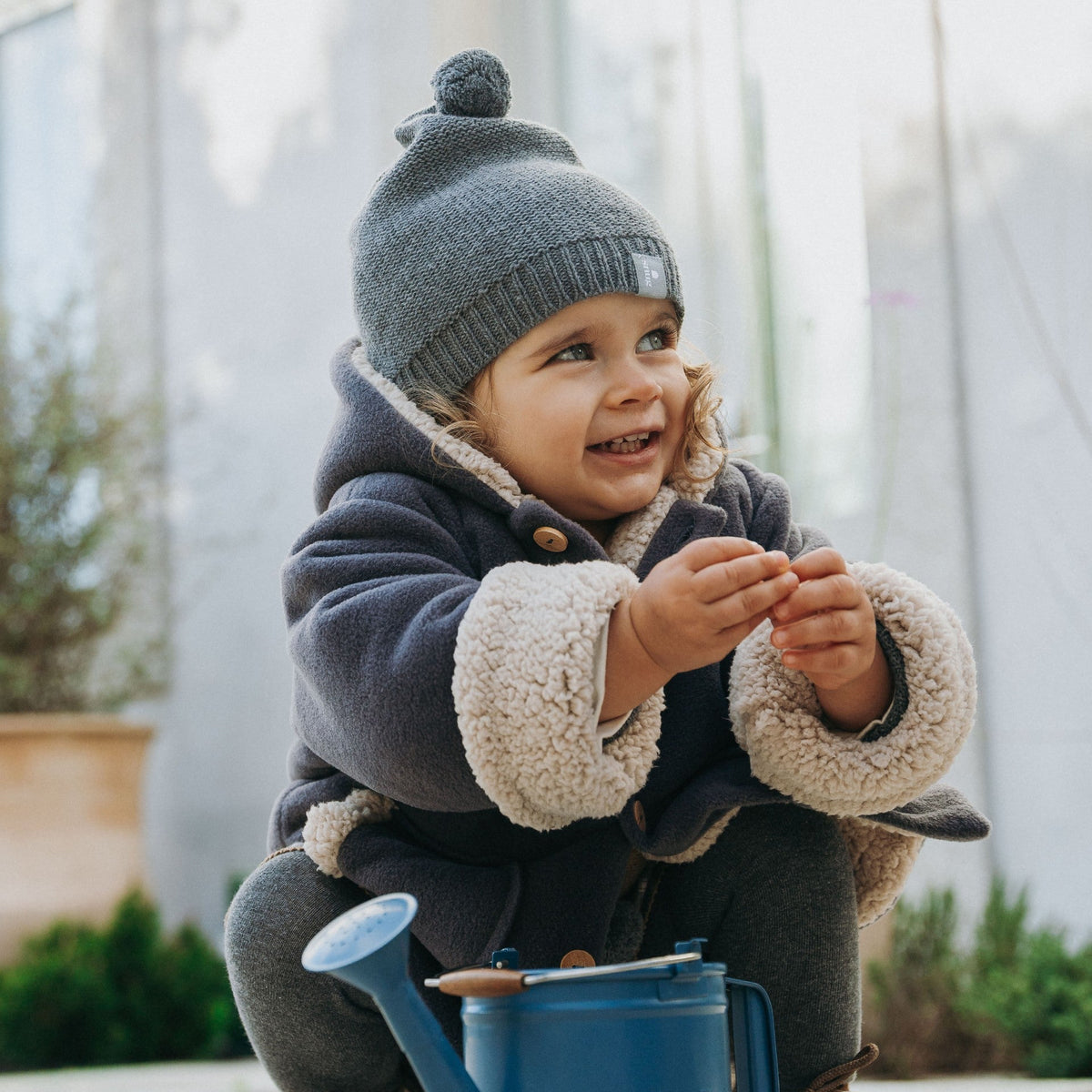 A toddler wearing a navy blue winter coat with beige sherpa lining, a gray knit beanie, and brown boots, crouching outdoors next to a blue watering can.