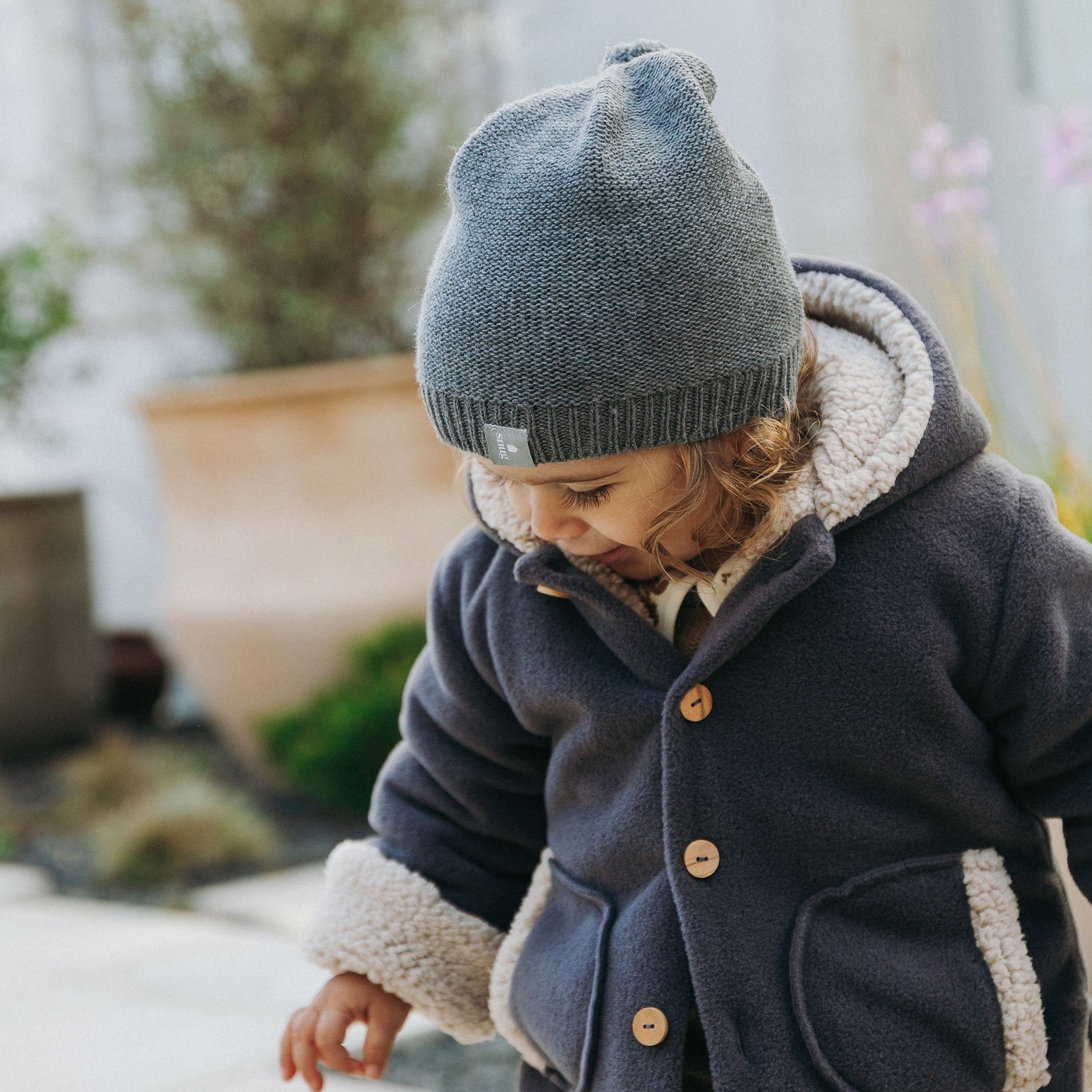 A toddler wearing a dusty pink knit beanie and cardigan over cream corduroy overalls, standing outdoors in a garden setting.