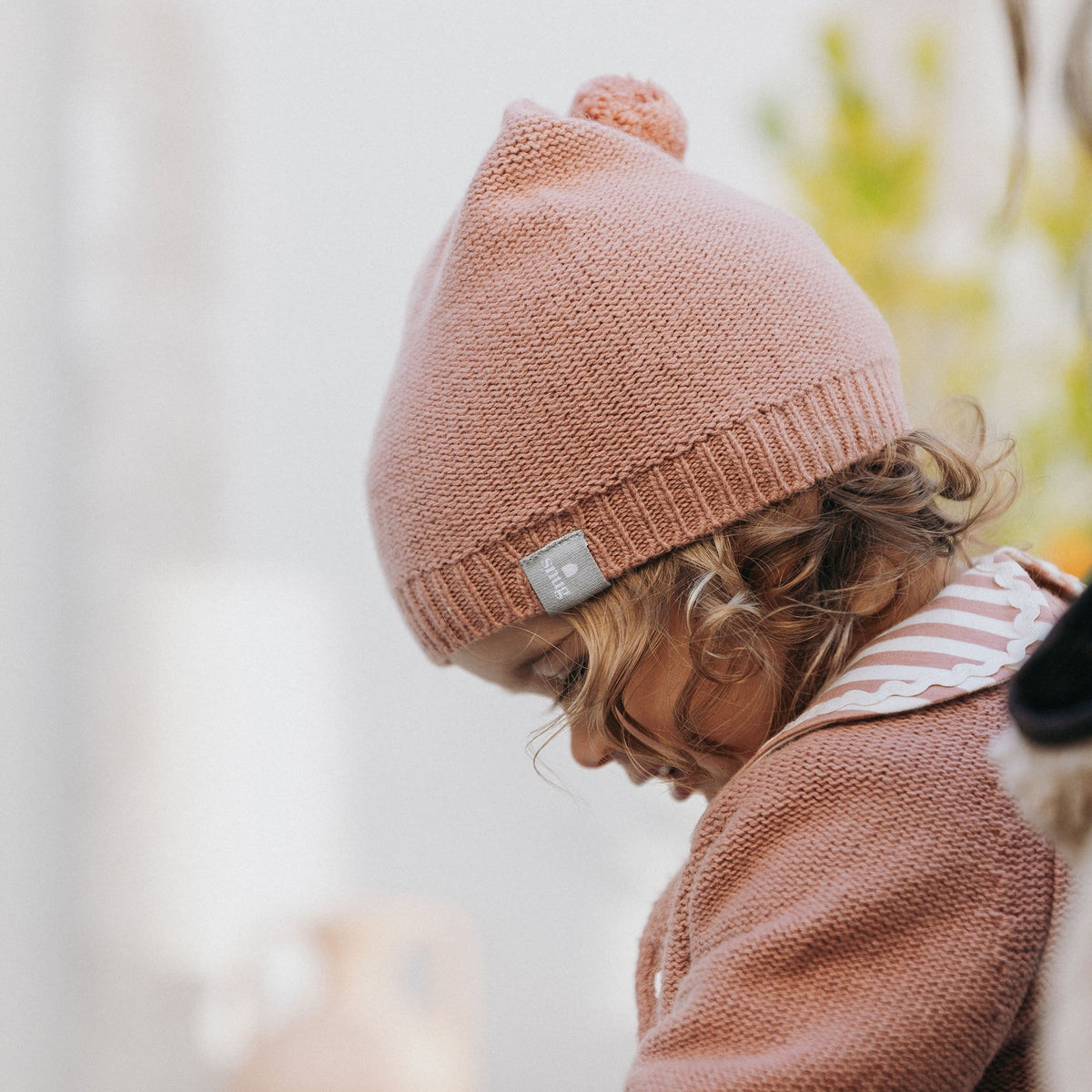 A child wearing a dusty rose knit beanie with a small silver brand tag and a matching knit sweater, shown in a softly blurred indoor setting.