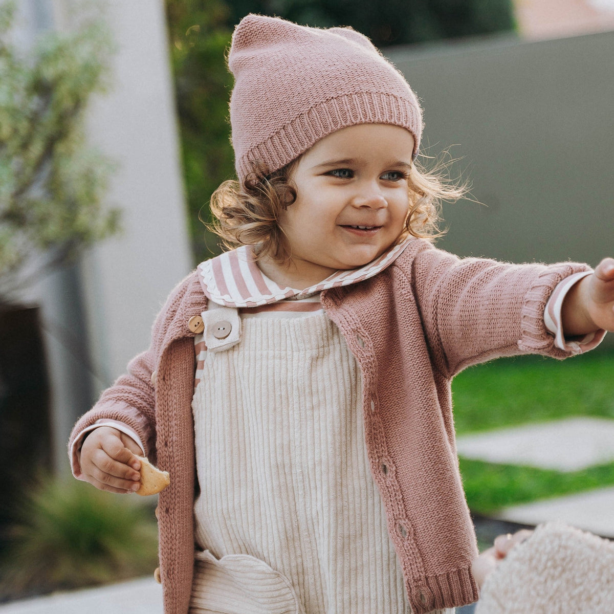 A toddler wearing a dusty pink knit beanie and cardigan over cream corduroy overalls, standing outdoors in a garden setting.
