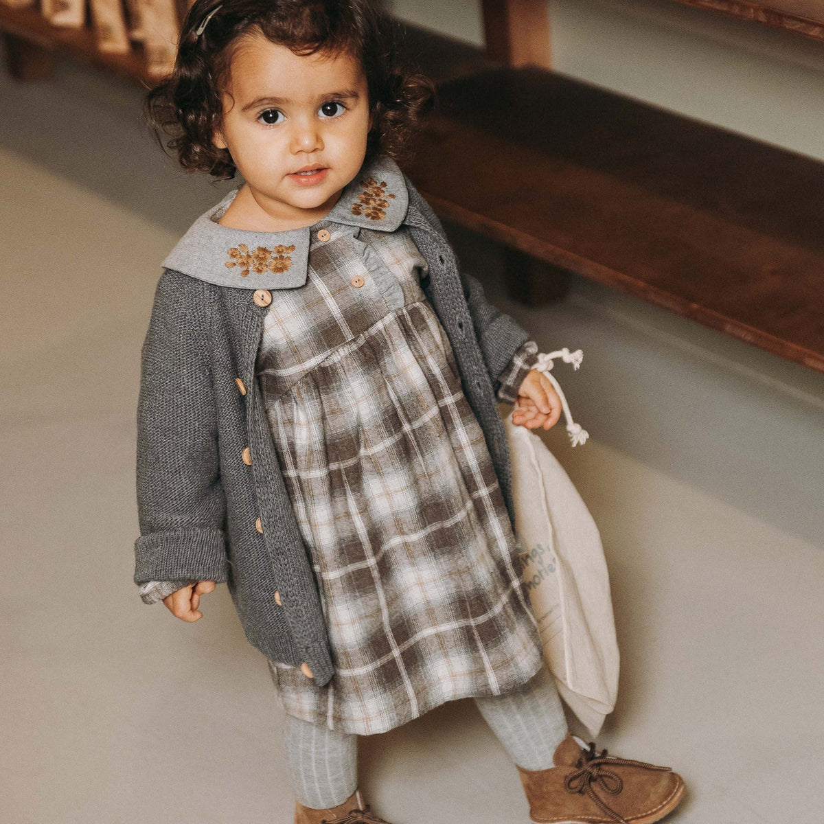 A toddler girl wearing a gray knit cardigan over a beige and gray plaid dress, paired with gray knit leggings and brown suede shoes, holding a cream drawstring bag in a boutique setting.