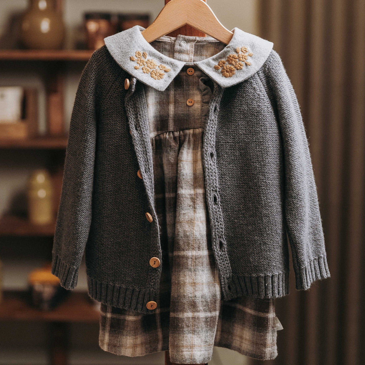 A children's gray knit cardigan with a light blue embroidered collar, layered over a brown and beige plaid dress, hanging on a wooden hanger.