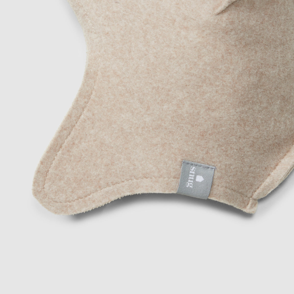 Close-up of a beige fleece baby hat  with a small gray fabric tag reading 'Snug' on a light gray background.
