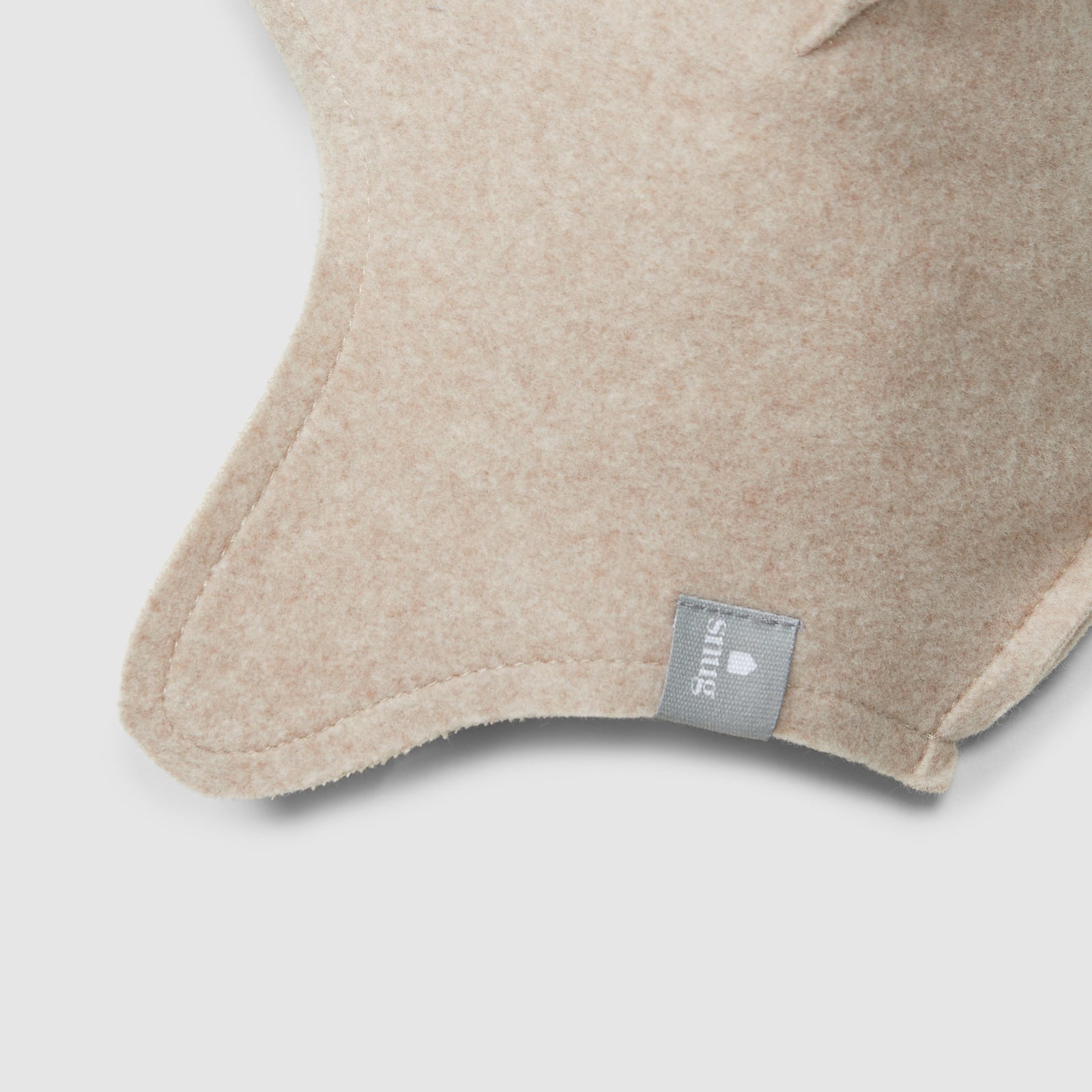 A light beige knit baby beanie hat with ear flaps and a small gray brand tag, shown on a neutral light gray background.