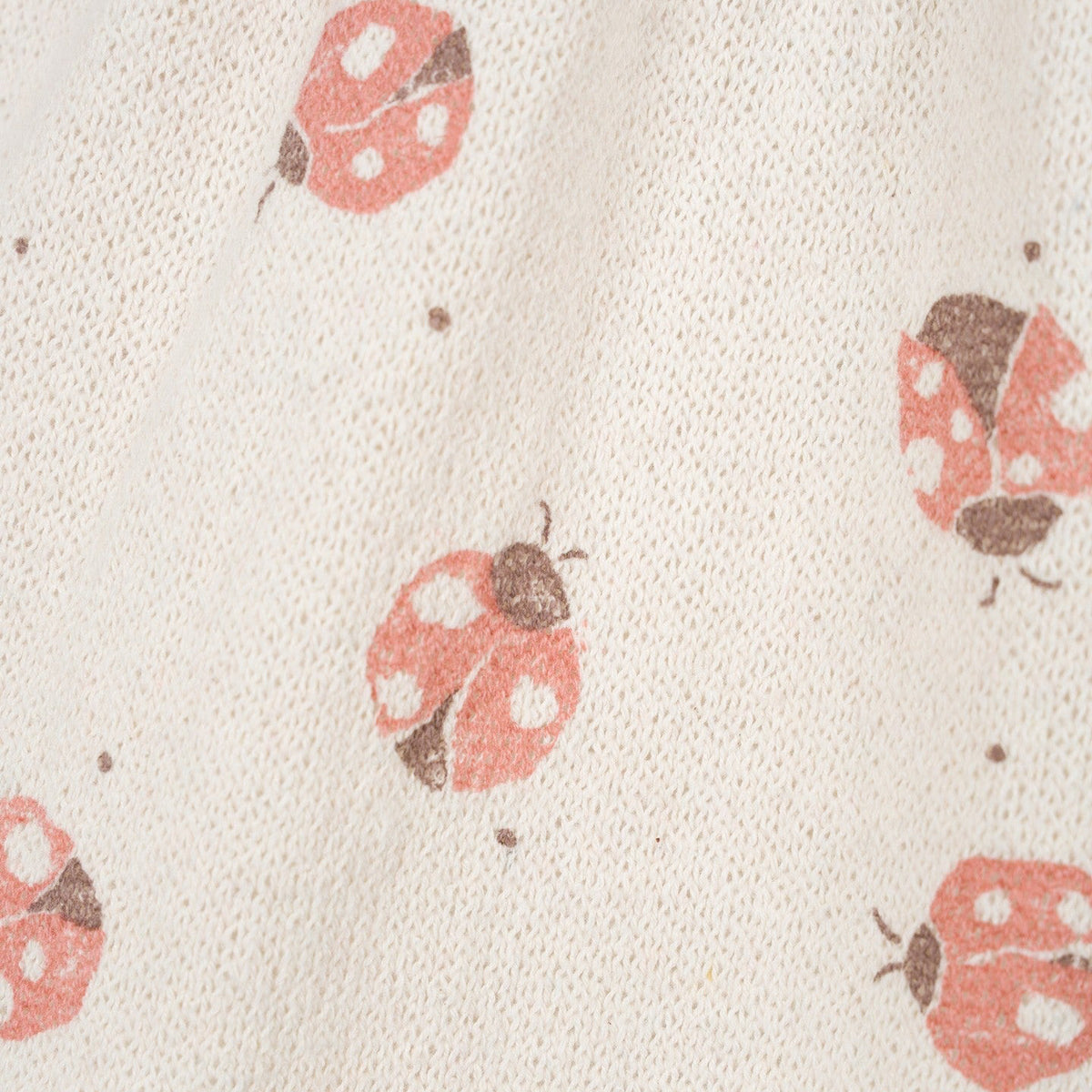Close-up of a textured cream knit fabric with a repeating pattern of pink ladybugs and small brown dots.