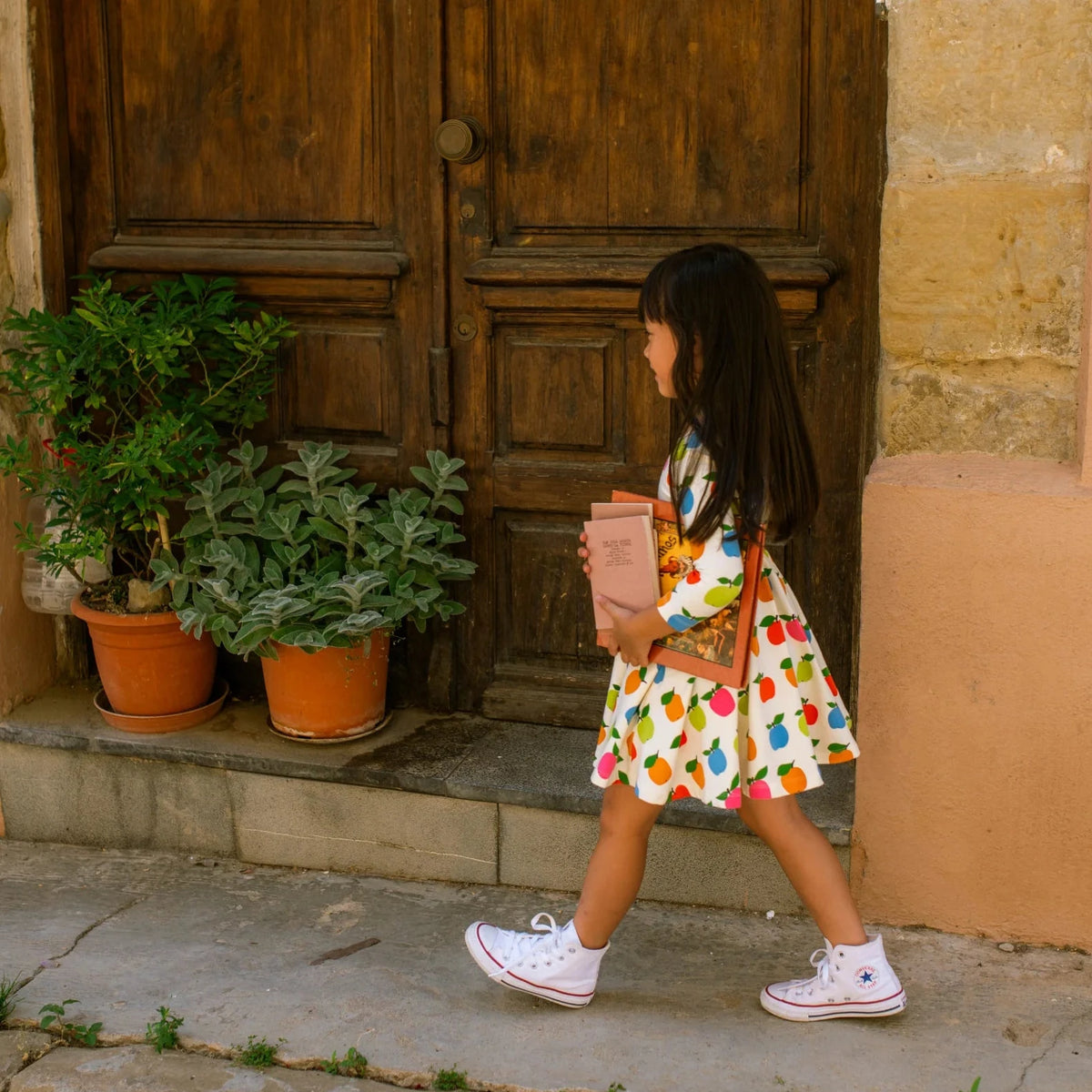 Young girl in a colorful apple print dress walking past a wooden door with potted plants.