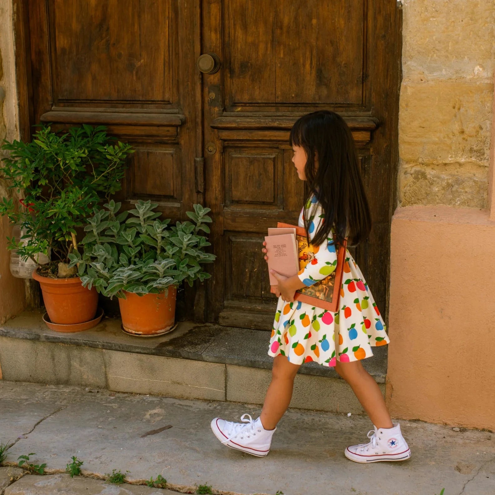 Young girl in a colorful apple print dress standing outside a wooden door.