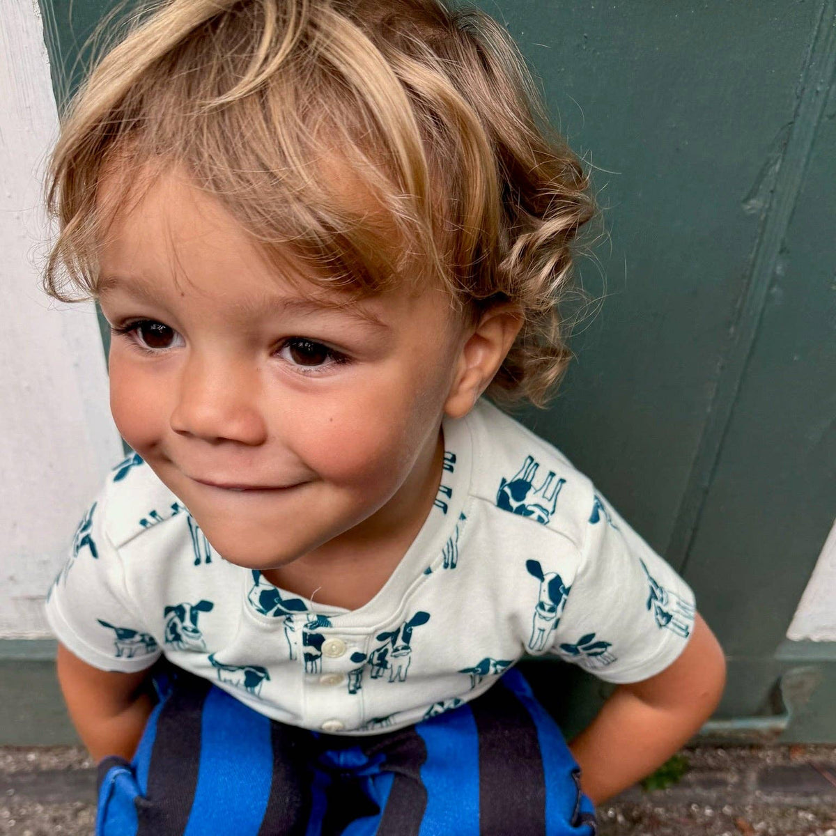 A young child crouching outdoors in front of a dark green door, wearing a white short-sleeve t-shirt with a blue Mickey Mouse pattern, blue and black striped pants, and blue patterned clogs.