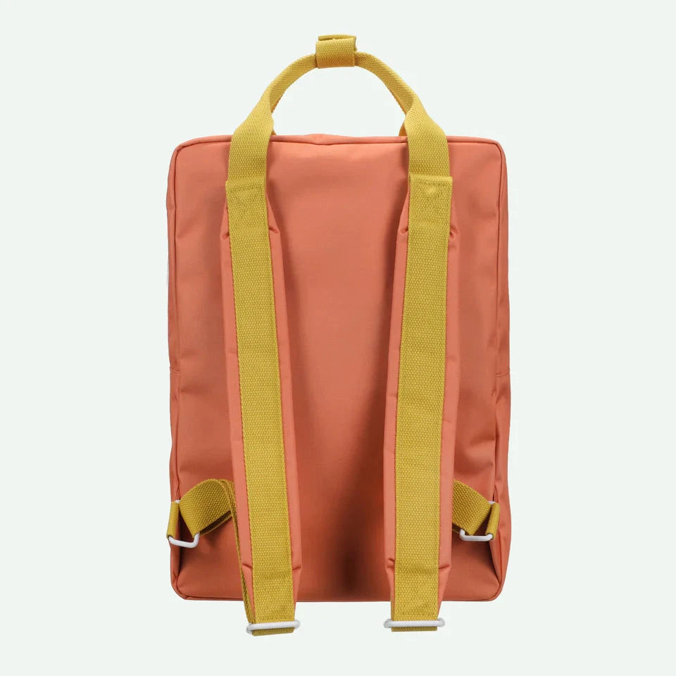 Person wearing an orange backpack with a green apple charm in a room with floral wallpaper.
