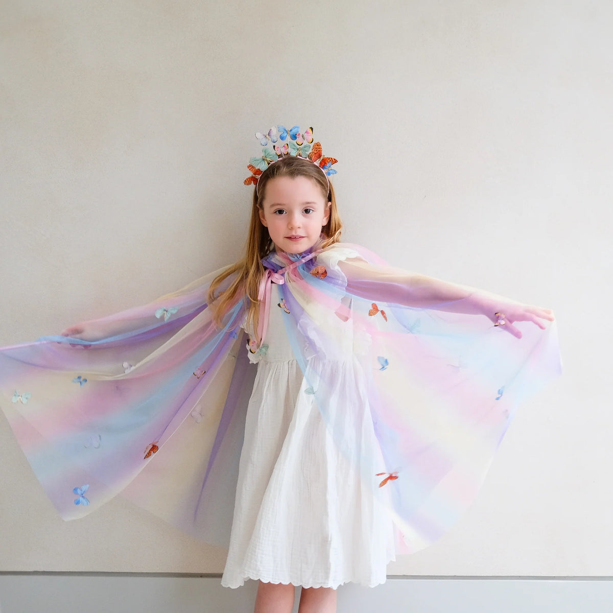 Young girl in a white dress with a colorful cape and crown against a plain background