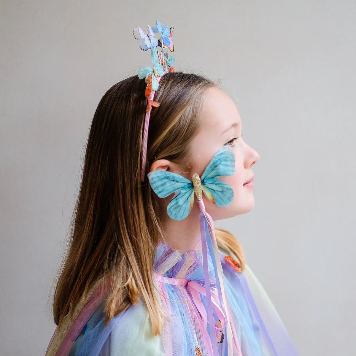 Child wearing colorful butterfly headband holding a butterfly wand against a plain background