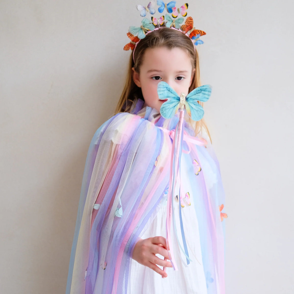 Child in a butterfly costume with a colorful headpiece and wand against a plain background