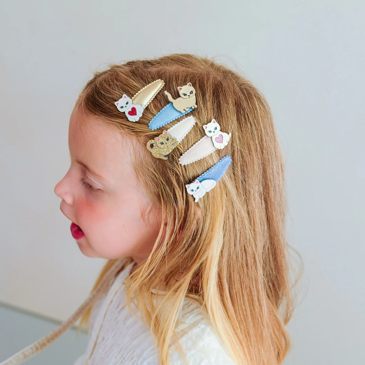 A side-profile view of a young girl with long blonde hair wearing six cute animal-themed hair clips in pastel colors, set against a plain light gray background.