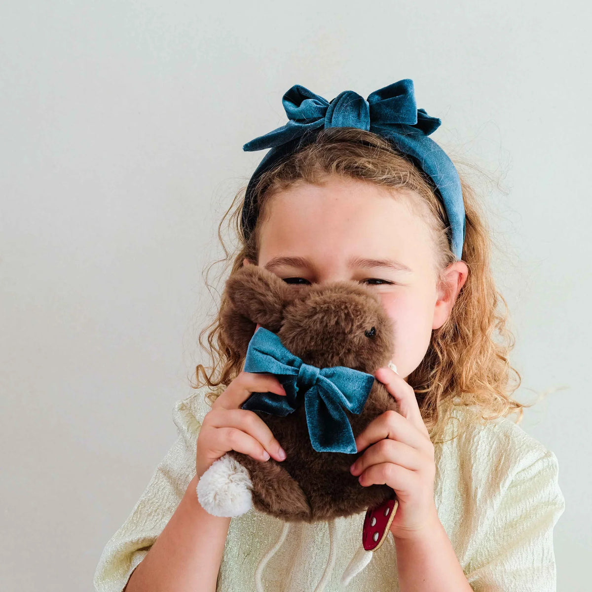 A young girl wearing a teal velvet bow headband, holding a brown plush bunny bag with a matching teal velvet bow against her face.