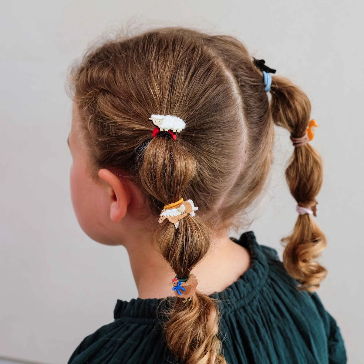 A child shown from the back wearing a dark green top, with light brown hair styled in two braided pigtails decorated with small colorful hair clips and elastic bands against a plain light gray background.