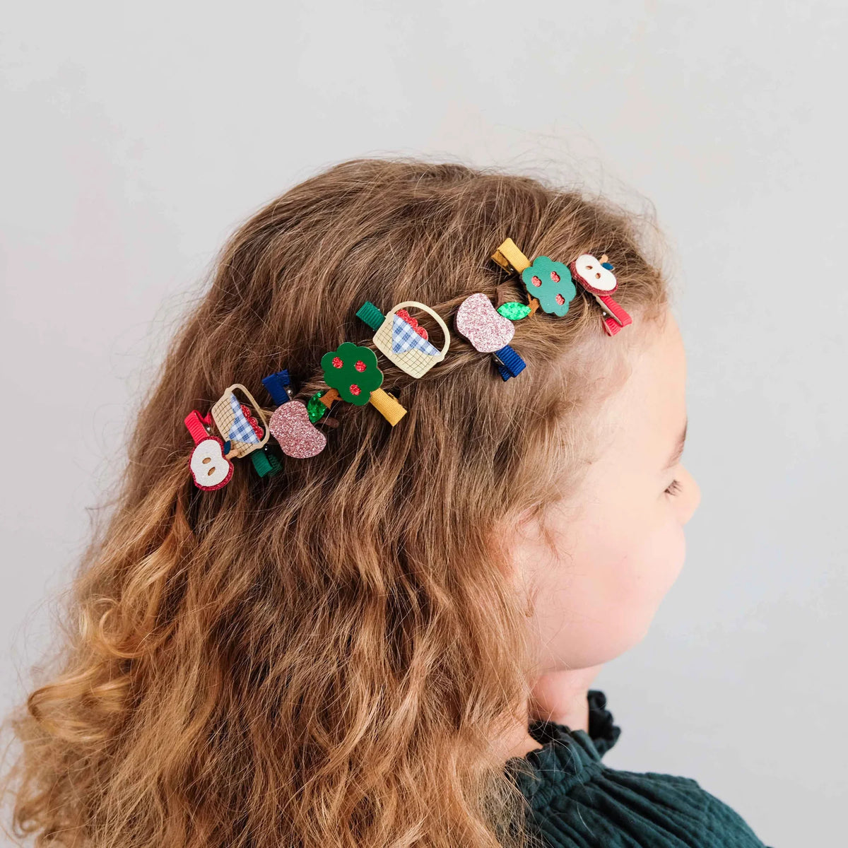 A side-profile view of a young girl with wavy light brown hair wearing a row of colorful decorative hair clips shaped like picnic items and fruit on a plain light gray background.
