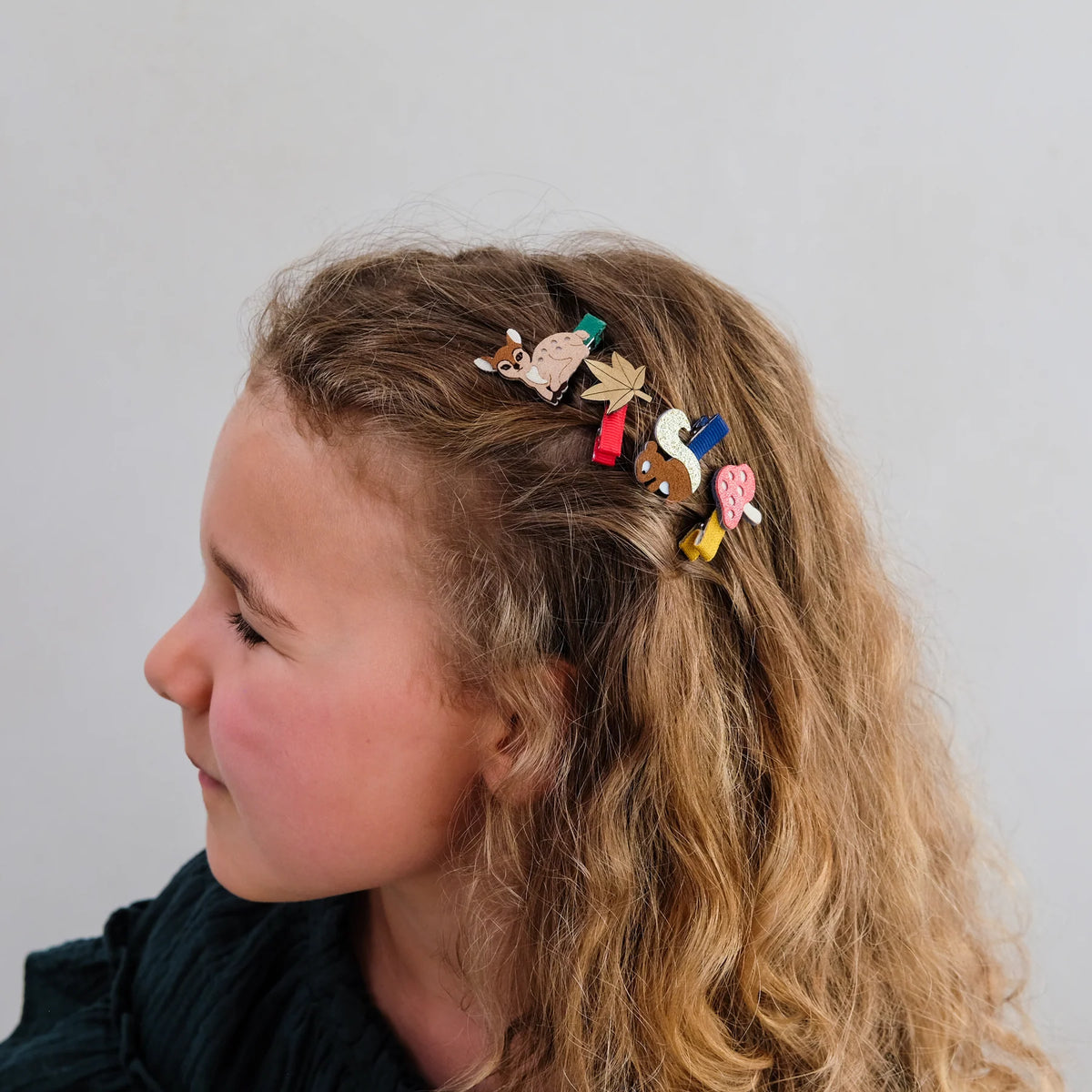 A side-profile view of a young girl with long, wavy light brown hair wearing several colorful wooden hair clips shaped like animals and objects, against a plain light gray background.