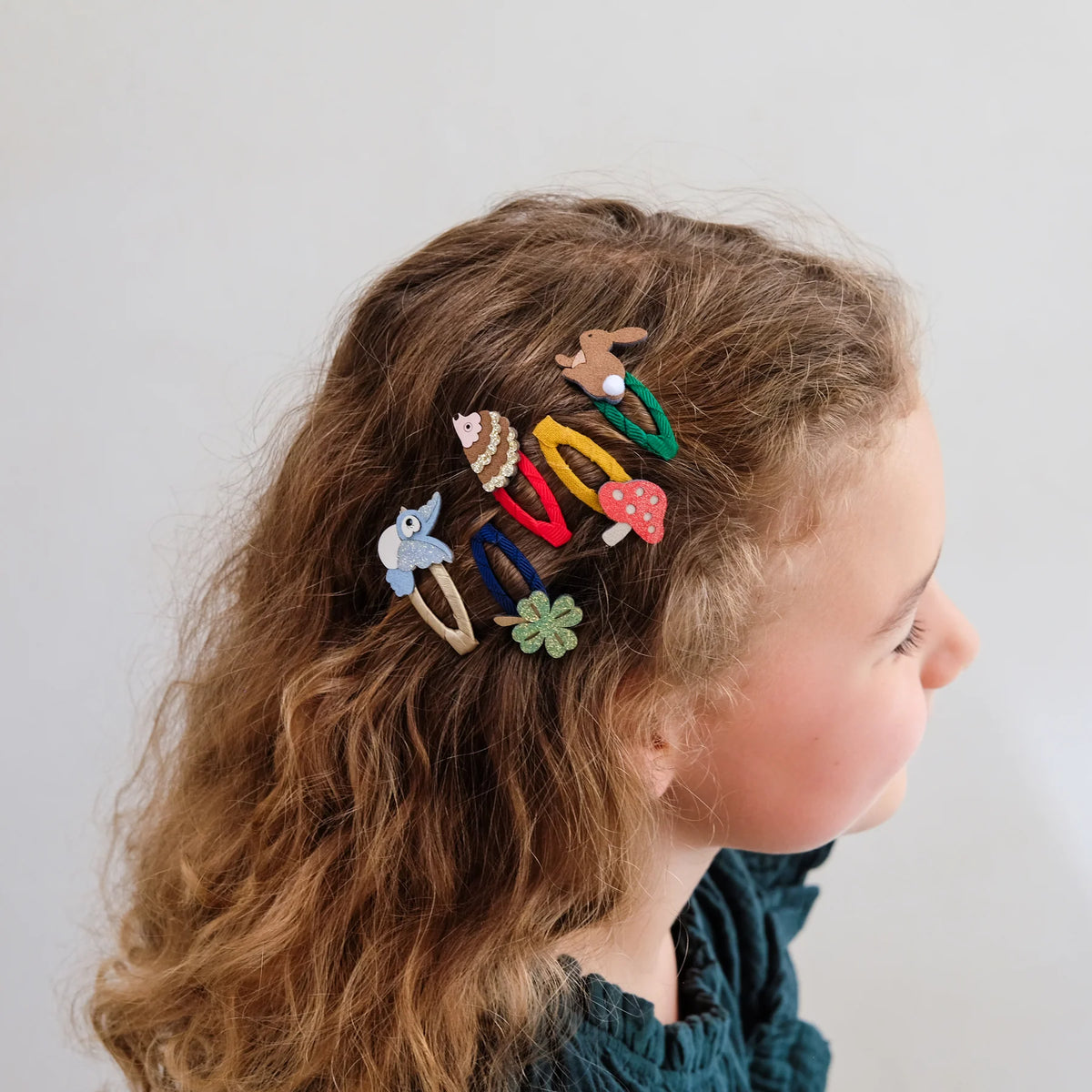 A side-profile view of a young girl with wavy light brown hair wearing several colorful woodland-themed hair clips against a plain light gray background.