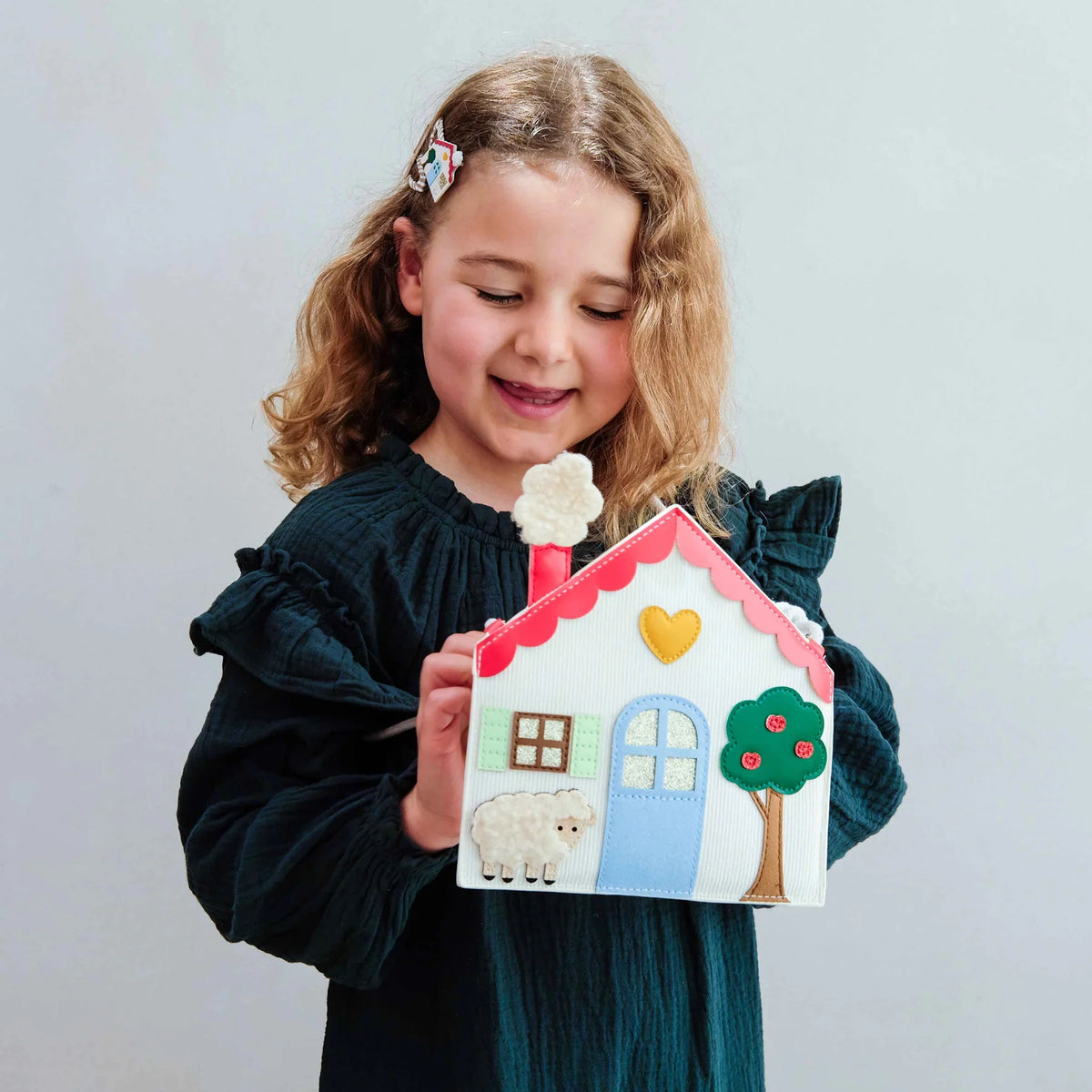 A young girl wearing a dark teal dress holds a handmade  house bag with a sheep, tree, and yellow heart, set against a plain light gray background.