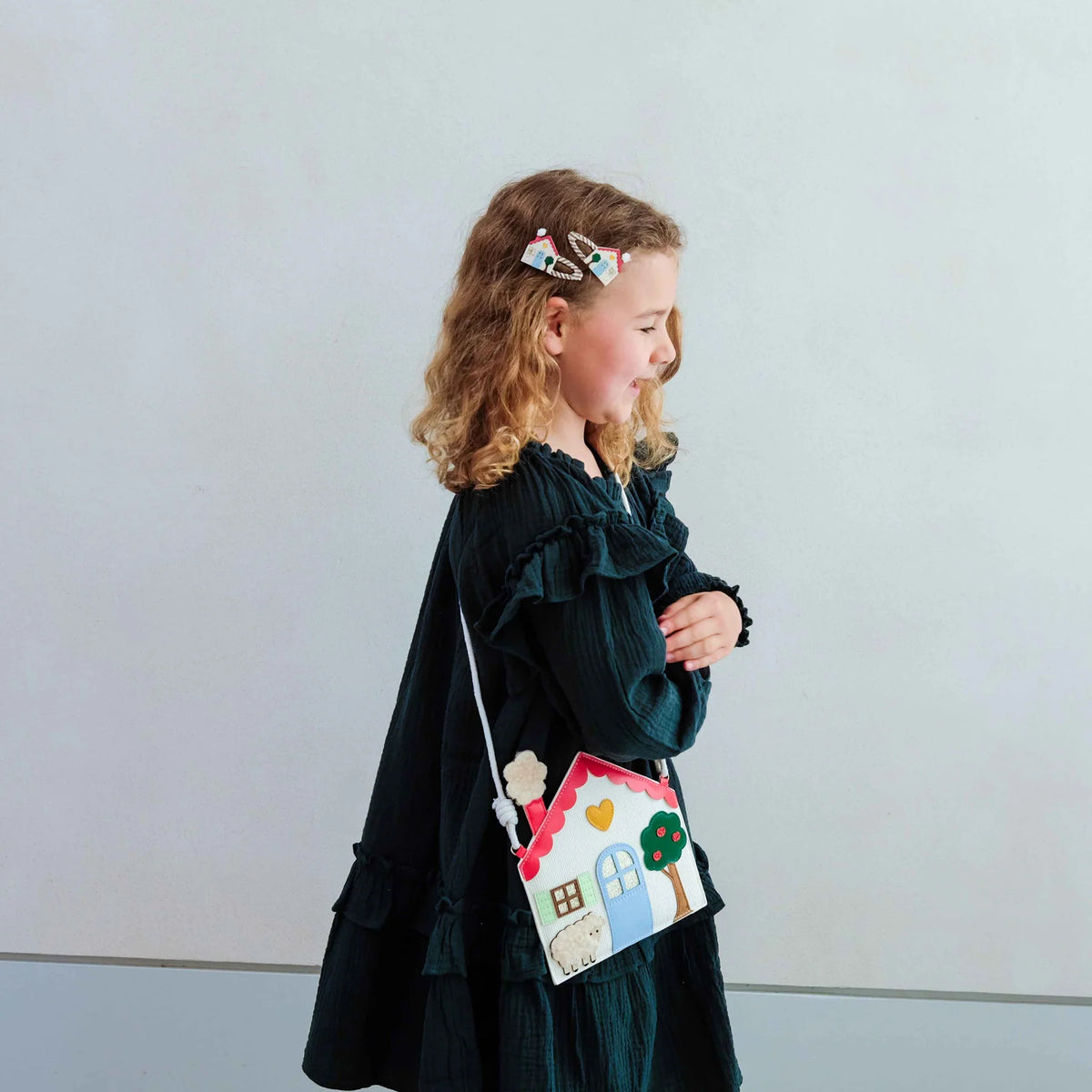 A young girl is shown in profile, wearing a dark green ruffled long-sleeve dress and carrying a small house-shaped crossbody bag with a colorful sheep and tree design. She is wearing a small hair clip.