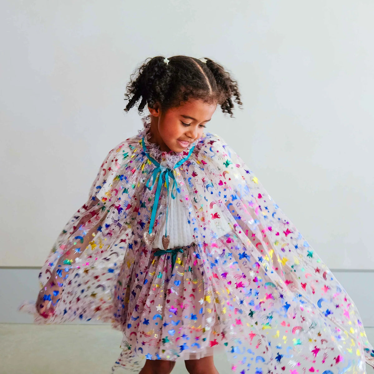 A young girl with curly hair wearing a sparkly, sheer cape and tutu with colorful star patterns and a blue ribbon tie, twirling in a bright indoor setting.