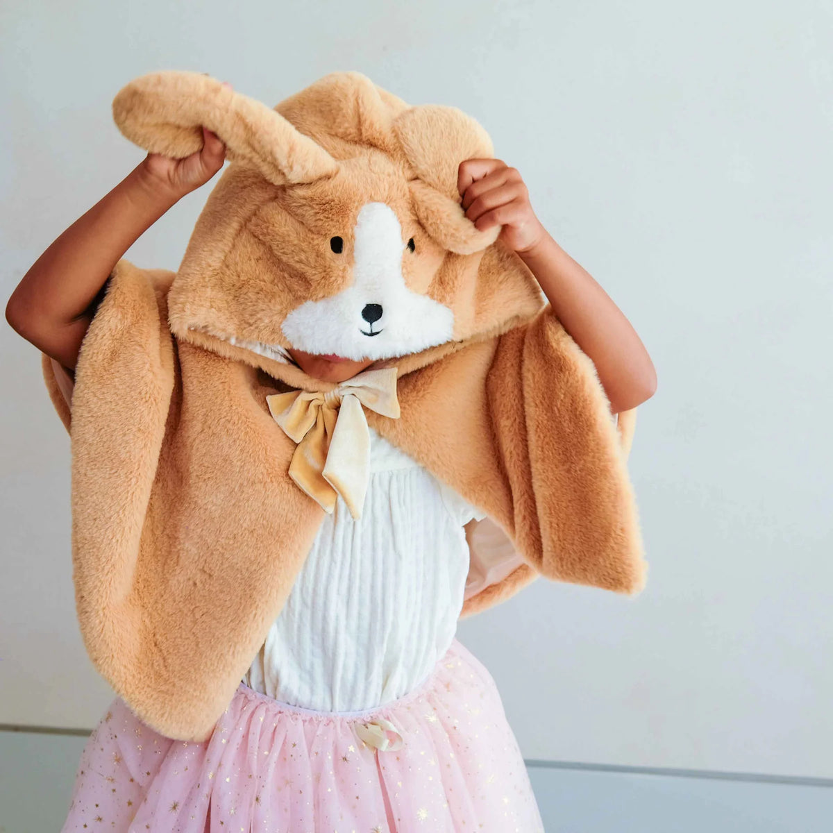 A child wearing a plush puppy hooded cape in a warm tan-brown color, featuring floppy ears, a white snout and forehead, black embroidered eyes and nose, and a pale yellow bow at the neck. 