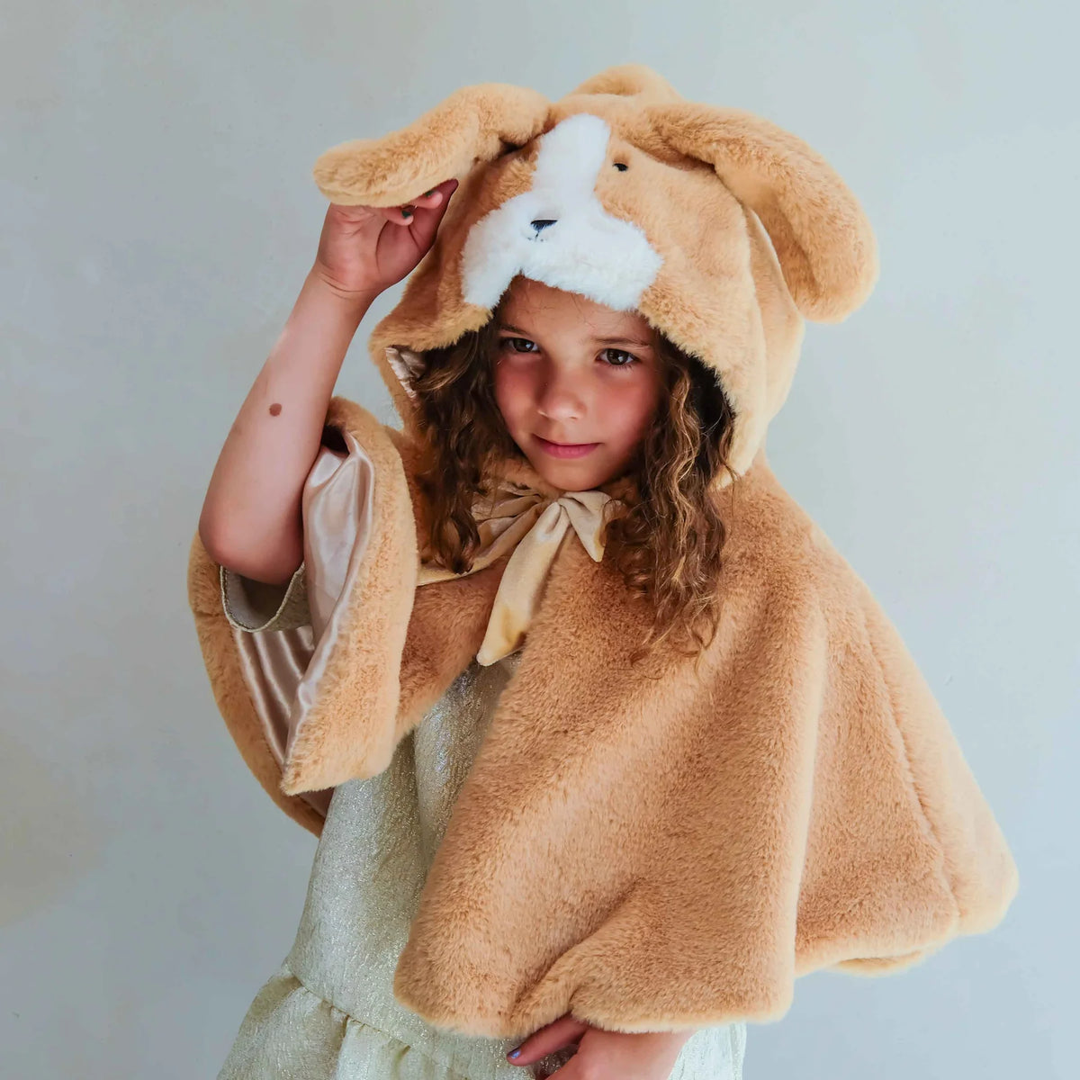 A young girl wearing a plush tan puppy hooded cape with floppy ears and a white snout detail, posed against a plain light gray background.